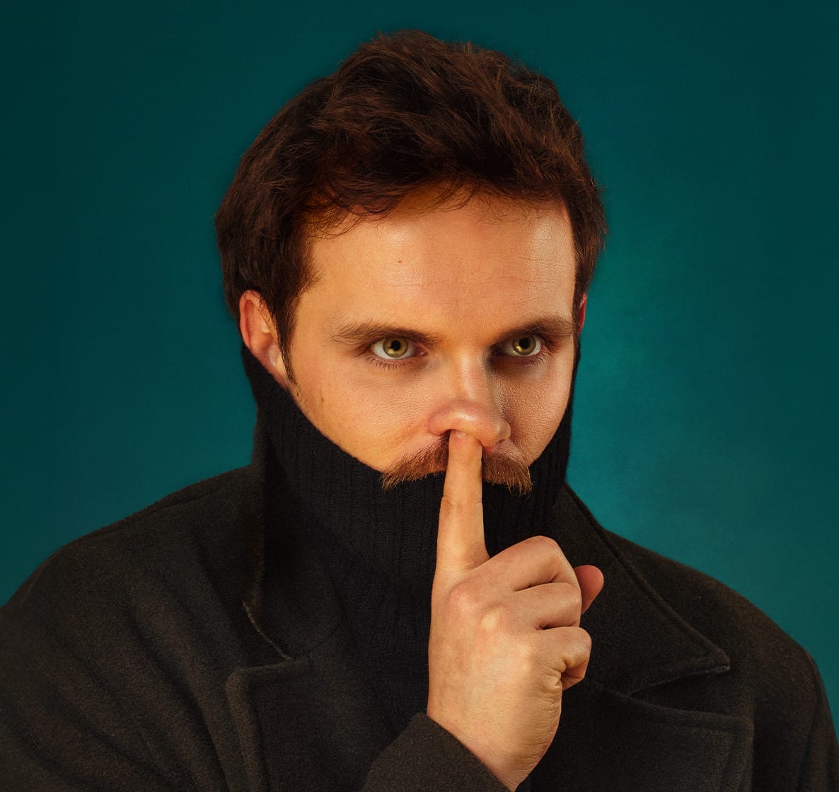 A man in a black turtleneck and moustache holds his finger up to his nose